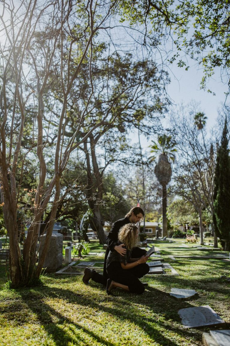 Two women embracing in a sunlit cemetery surrounded by trees.